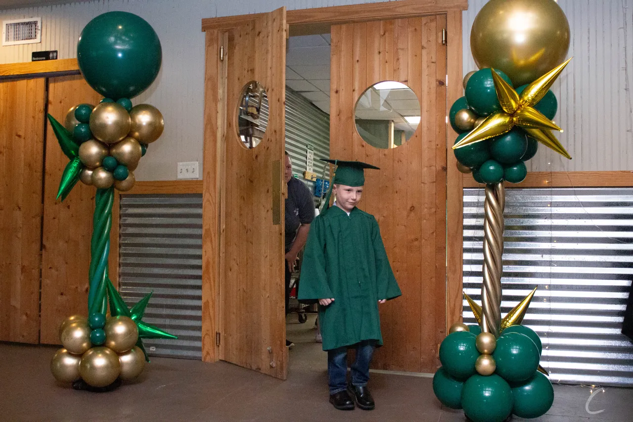 A young graduate walking through wooden doors between celebratory balloon columns