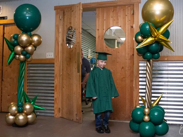 A young graduate walking through wooden doors between celebratory balloon columns