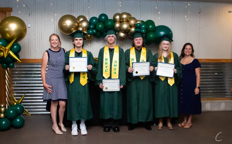 NHTC's graduating class in green caps and gowns holding diplomas in front of a balloon arch