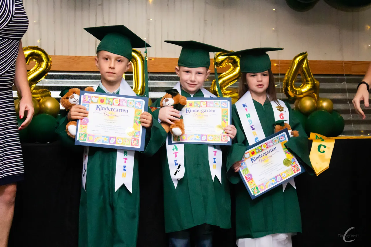 Three kindergarten graduates in tiny caps and gowns holding certificates