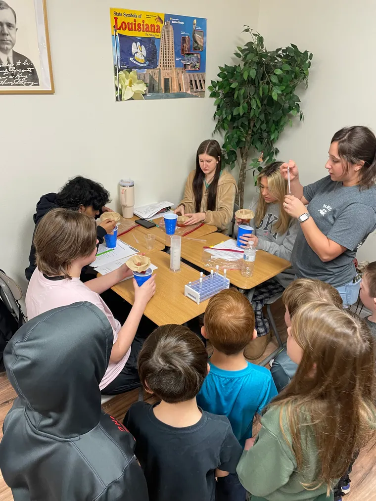 A mixed-age science class gathered around a table for an experiment, with a Louisiana poster on the wall