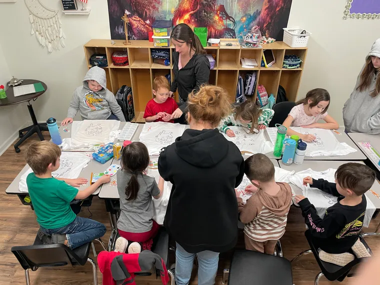A teacher helping students decorate t-shirts at a long classroom table