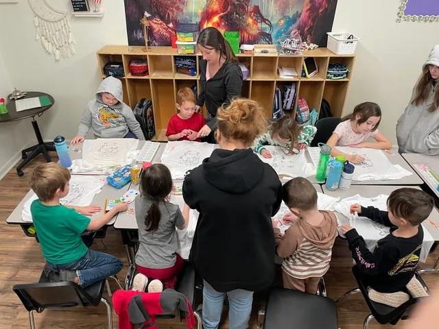 A teacher helping students decorate t-shirts at a long classroom table